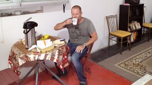 A man sitting at a table with a kettle, a mug, and some food items. There is a chair and a bookshelf in the background. - HOSPEDERIA Calle 5 (Callao)