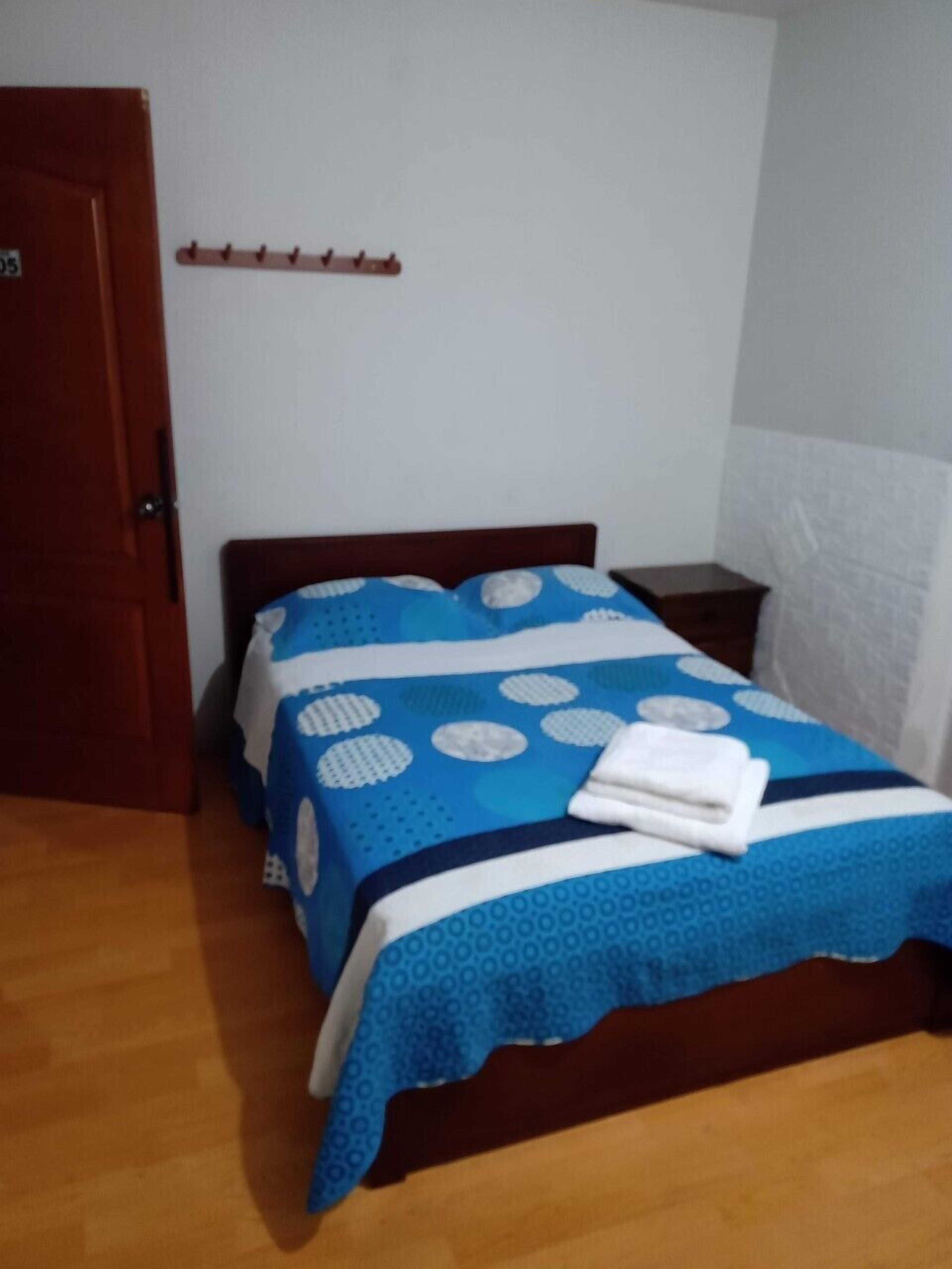 Un dormitorio con una cama de madera, una colcha de cama azul y blanca, una toalla blanca sobre la cama, una mesita de noche de madera y una puerta de madera.
