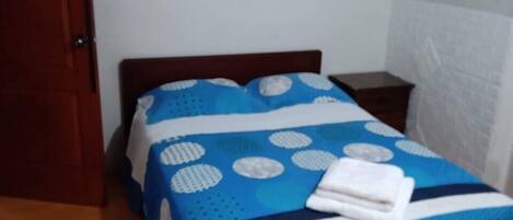 Un dormitorio con una cama de madera, una colcha de cama azul y blanca, una toalla blanca sobre la cama, una mesita de noche de madera y una puerta de madera.