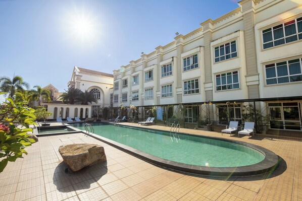 Uma piscina com espreguiçadeiras, um elemento decorativo rochoso e um edifício com várias janelas e varandas.