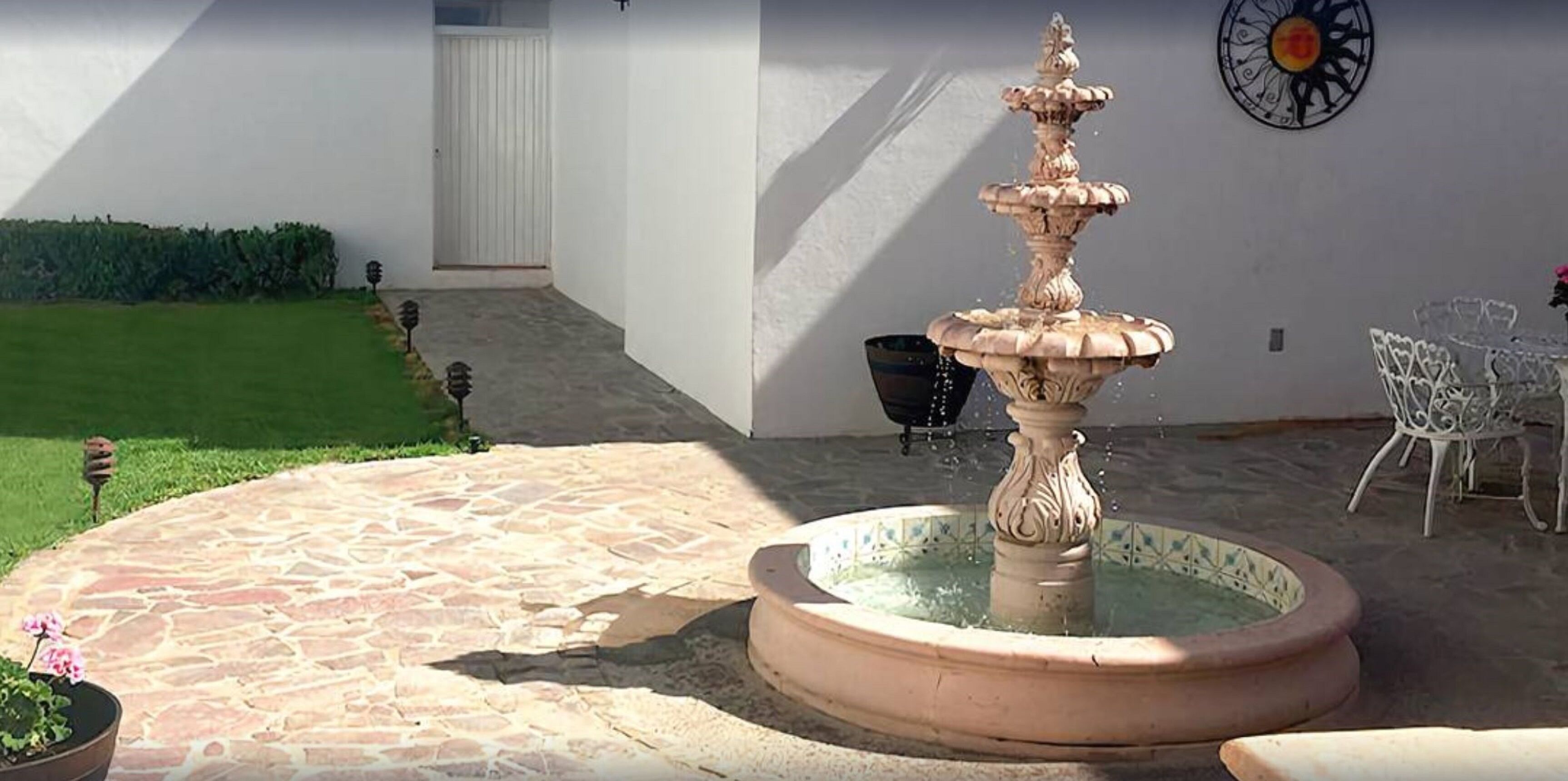 Un patio de piedra con una fuente en el centro, sillas blancas y una pared de color blanco con un detalle metálico decorativo.