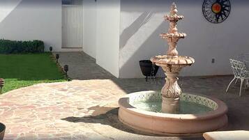 Un patio de piedra con una fuente en el centro, sillas blancas y una pared de color blanco con un detalle metálico decorativo.