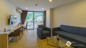 A modern living room with a sofa, armchair, and a glass coffee table. There is a desk with a computer and a TV mounted on the wall. - New Square Patong Hotel (Patong)