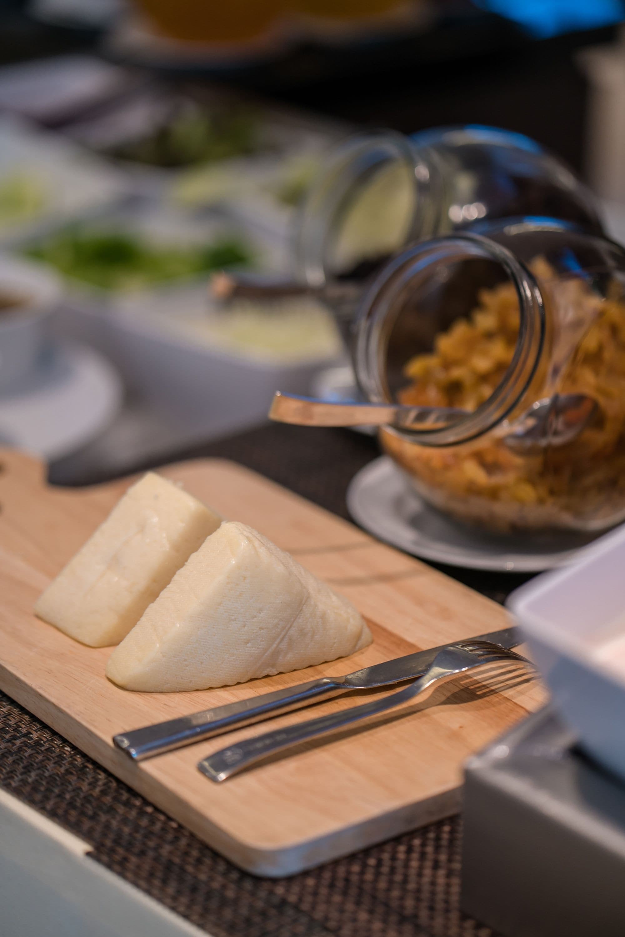 A wooden cutting board with two white items, a glass jar with a lid, and a metal fork and knife on a table.