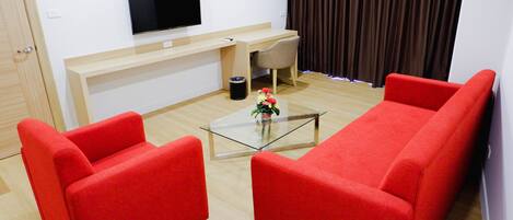 A modern living room with a flat-screen TV, a wooden desk, a glass coffee table, and two red sofas.