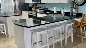 A modern kitchen with white cabinets, a stainless steel refrigerator, a microwave, and a sink. There are three white bar stools and a wooden chair. - 2000sq ft Villa 401, Ocean View steps to the beach, small pool hot tub 4 king bd (North Myrtle Beach)