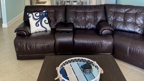 A living room with a dark brown leather sectional sofa, a round coffee table with a blue and white design, and a kitchen in the background with white cabinets and a refrigerator. - 2000sq ft Villa 401, Ocean View steps to the beach, small pool hot tub 4 king bd (North Myrtle Beach)