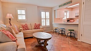A living room with a sofa, a coffee table, and a lamp. There are two windows and a kitchen area with a counter and stools. - Sunseeker - Xanadu by Coastal Dreamin (Santa Rosa Beach)