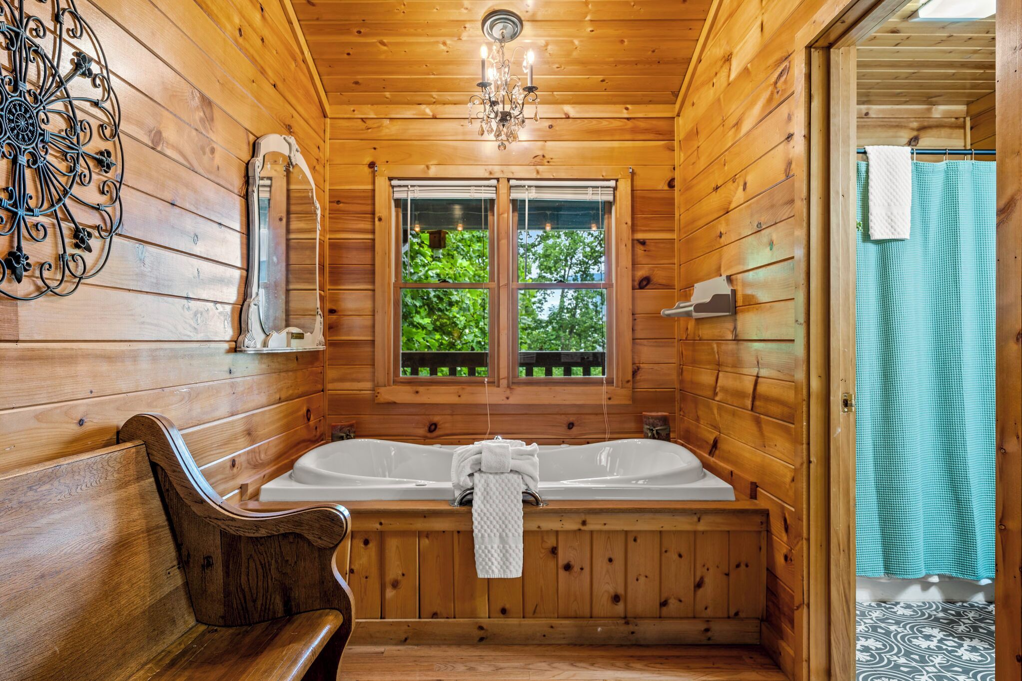 A wooden cabin bathroom with a large bathtub, wooden benches, a chandelier, and a window with a view of greenery.