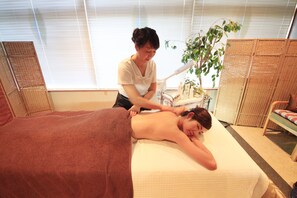 A massage therapy room with a massage table, a client receiving a back massage, a massage chair, and a plant in the background.