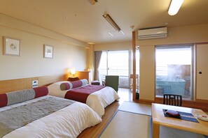 A hotel room with two beds, a table with a chair, a window with a view, and an air conditioning unit.