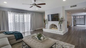 Amplia sala de estar con chimenea, ventilador de techo y un ventanal con cortinas translúcidas.