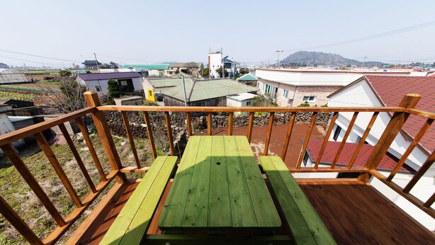 Een houten terras met een groene picknicktafel, omringd door een houten balustrade, met uitzicht op een woongebied met huizen en een heuvel in de verte.