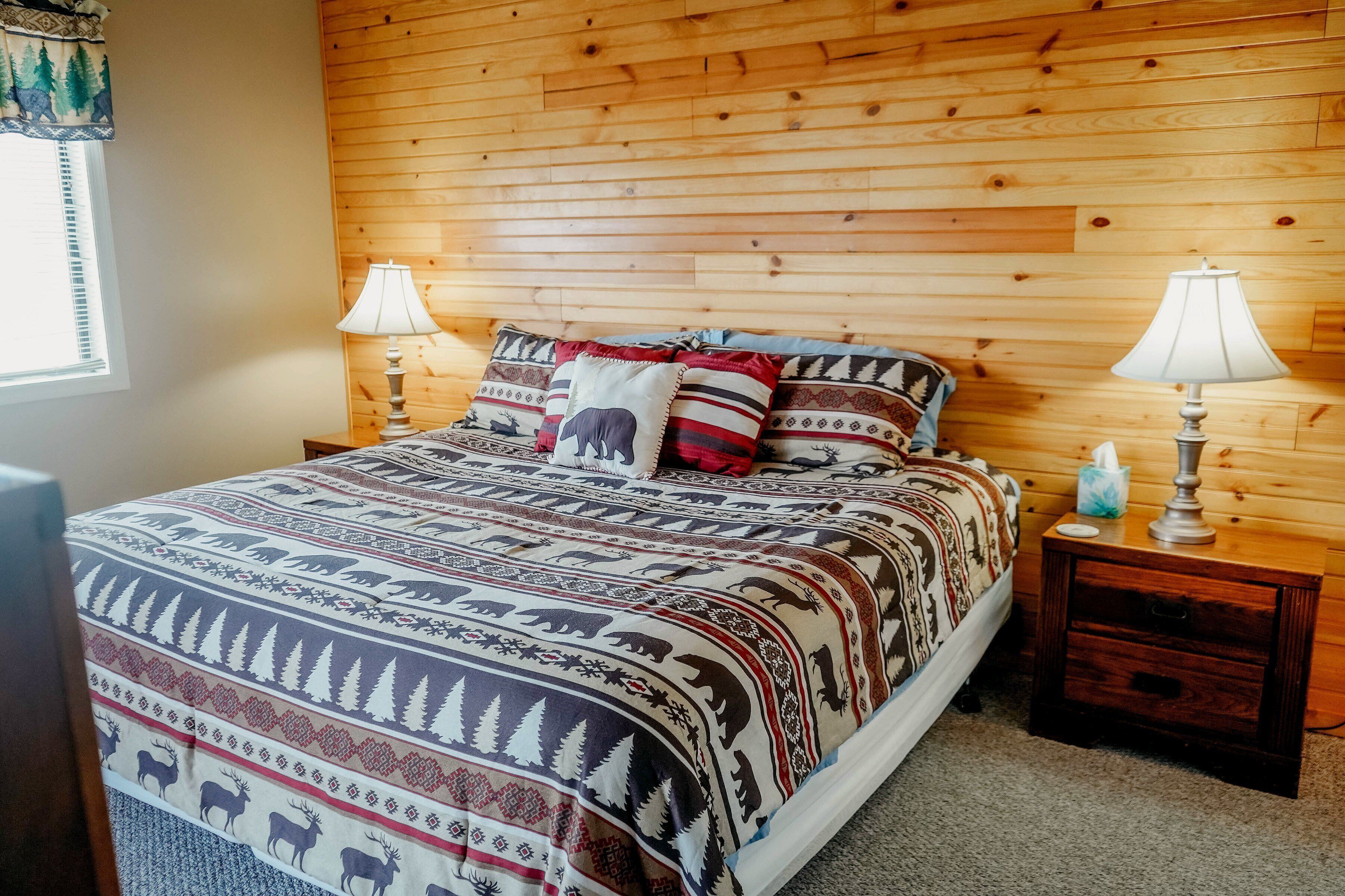 Een slaapkamer met een houten hoofdbord, een bed met een decoratieve spreidek en twee nachtkastjes met lampen, en een raam met jaloezieën.