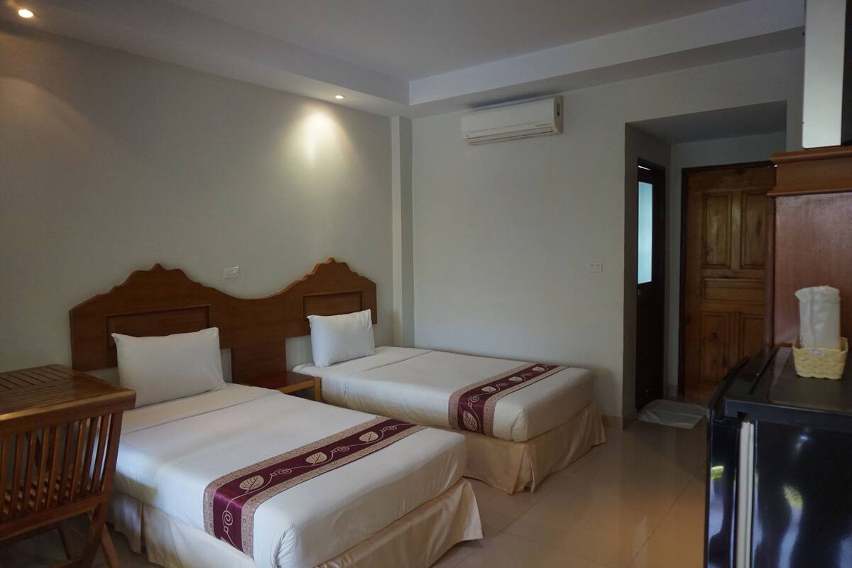 Quarto de hotel com duas camas, cabeceira de madeira, uma mesa pequena e um frigorífico.