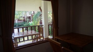 A room with a wooden table and chairs, a window with curtains, and a view of a garden with trees and a fence. - Buppha Resort (Takua Pa)