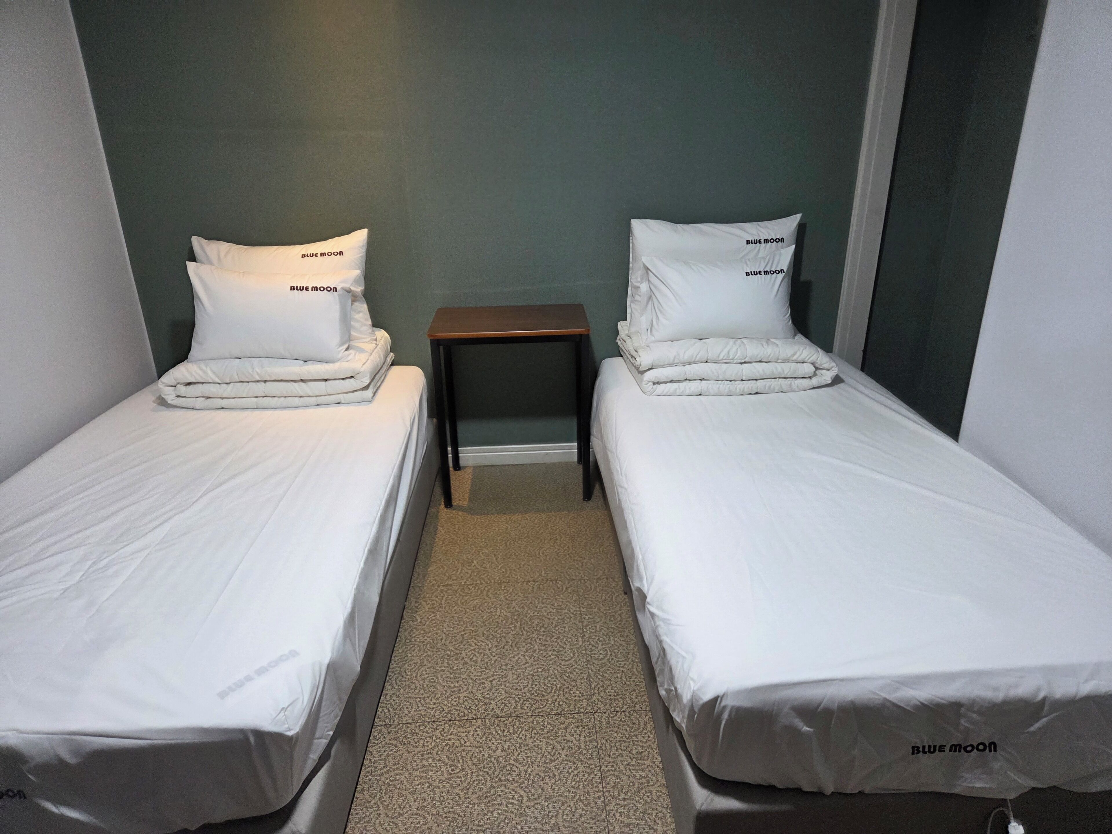 Dos camas individuales con ropa de cama y almohadas blancas, una mesita de madera pequeña entre ellas, y piso cubierto de alfombra.