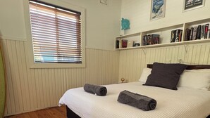 Ein Schlafzimmer mit Holzboden, einem Bett mit weißer Bettwäsche und zwei Kissen, einem Fenster mit Jalousien und einem Bücherregal mit Büchern und Dekoration.