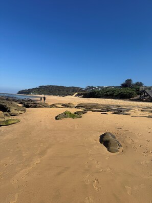 Uma praia arenosa com pegadas, um afloramento rochoso e um céu azul e límpido.