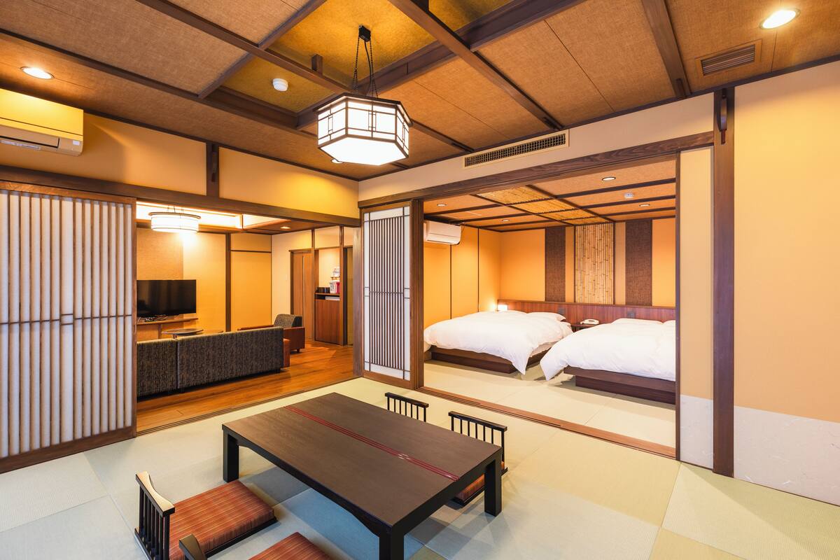 A traditional Japanese-style room with tatami flooring, a low table with cushions, a bed with white bedding, and a television in the corner.