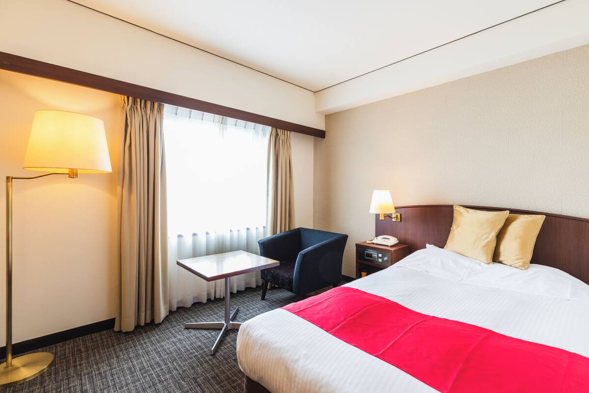 A hotel room with a bed, a desk, a chair, a lamp, and a nightstand.