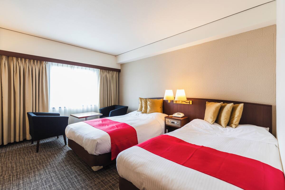 A hotel room with two beds, a desk, a chair, and a window with curtains.