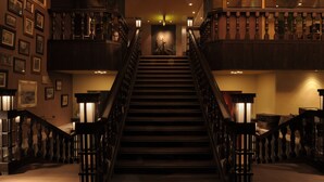 A grand staircase with ornate railings and wall-mounted lamps, leading to a second-floor landing with a view of the interior. - Urban Villa Konaya Hotel (Kofu)