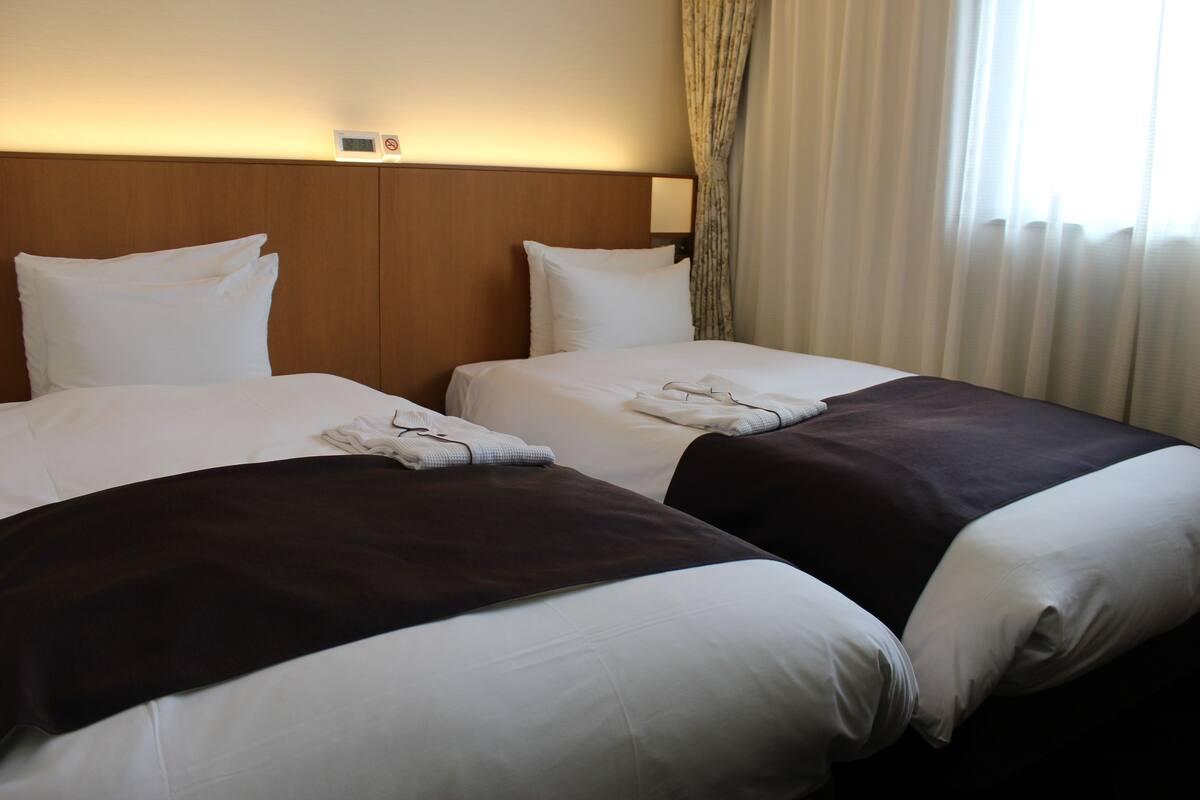A hotel room with two beds, each with white linens and a dark-colored throw blanket. There is a wooden headboard, a wall-mounted control panel, and a window with curtains.
