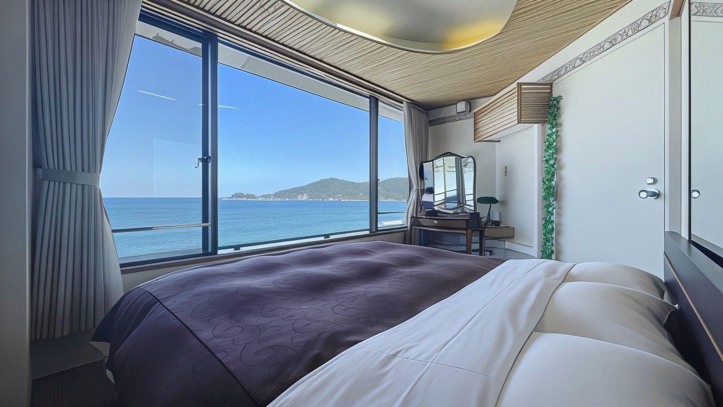 A modern bedroom with a large bed, a desk, and a view of the sea.