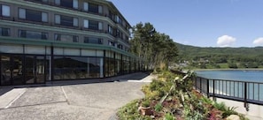 A multi-story hotel with a glass facade, a paved terrace, and a view of a body of water and mountains. - Sanriku Hana Hotel Hamagiku (Otsuchi-cho)