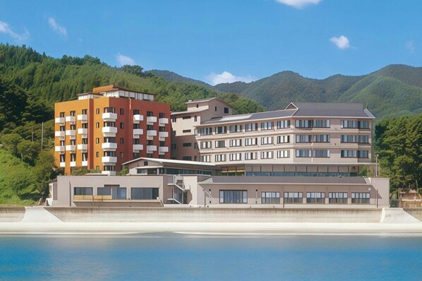 A multi-story hotel with balconies, a flat roof, and a view of a body of water and hills. - Sanriku Hana Hotel Hamagiku (Otsuchi-cho)