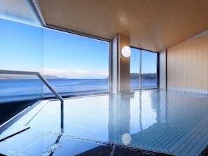 An indoor swimming pool with a view of the sea, a wooden ceiling, and a tiled floor.