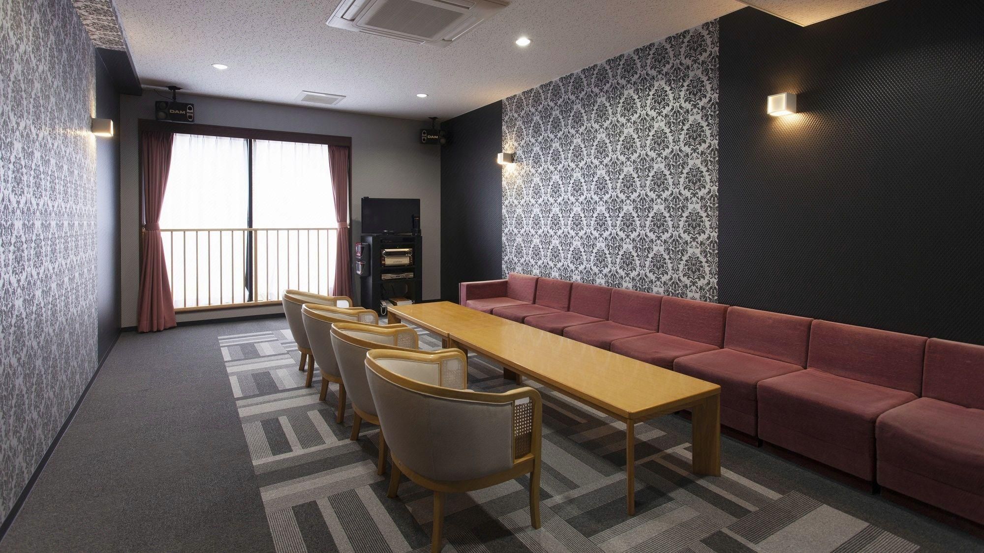 A conference room with a long table, chairs, a sofa, patterned wallpaper, and a window with curtains.