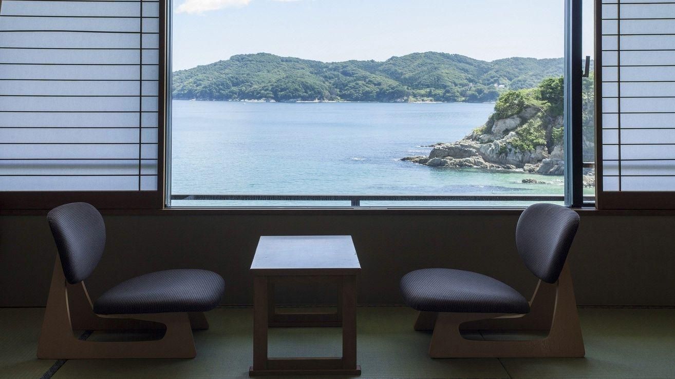 A room with tatami flooring, two chairs, a small table, and a view of a coastal landscape through a large window.