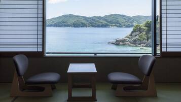 A room with tatami flooring, two chairs, a small table, and a view of a coastal landscape through a large window.