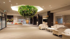 Un hall d’entrée spacieux agrémenté d’une composition florale suspendue au centre, d’un coin salon avec des canapés et une table basse, ainsi que d’un comptoir de réception orné d’une décoration colorée.