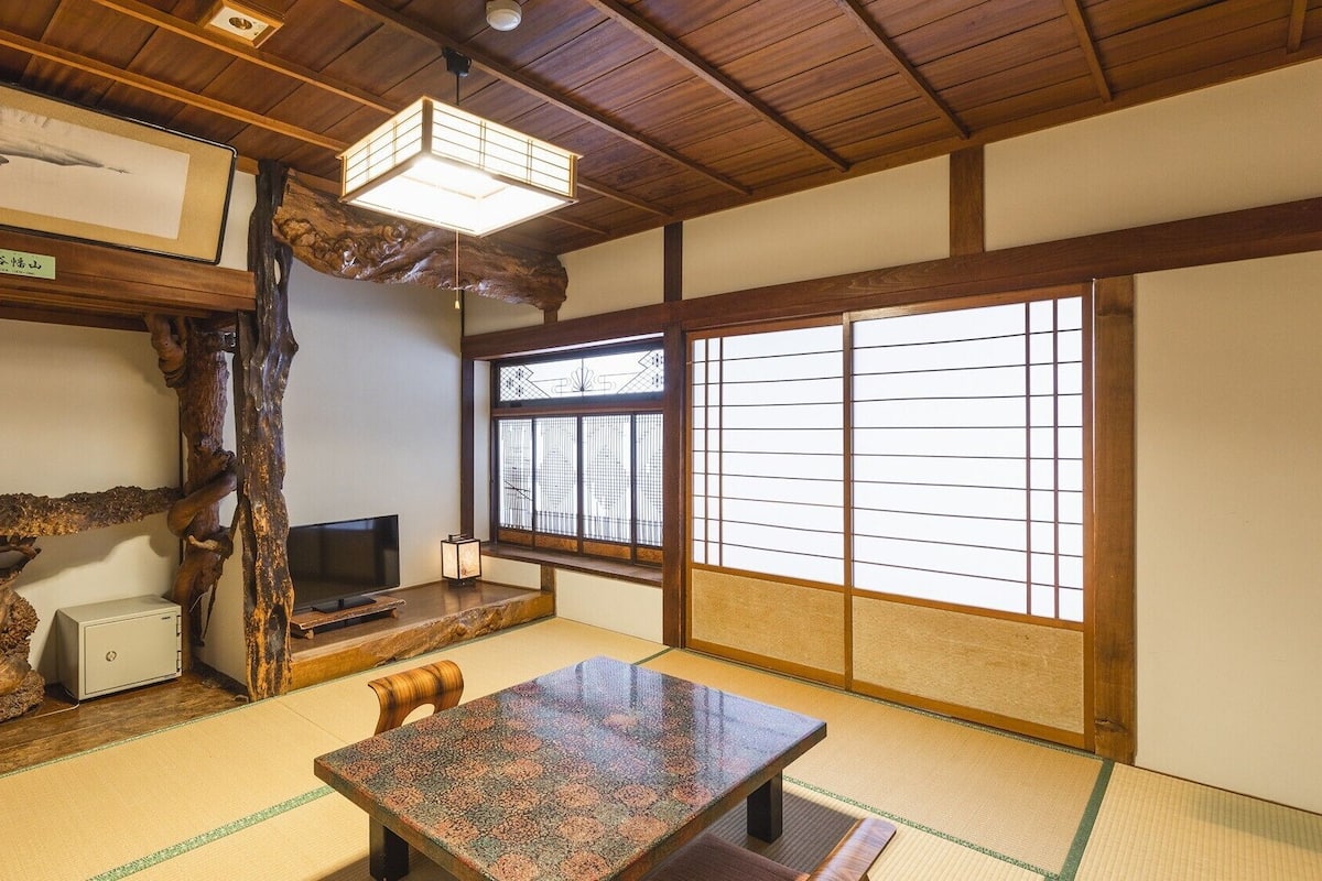 A traditional Japanese room with tatami flooring, a low table, a TV, and a large window with shoji screens.