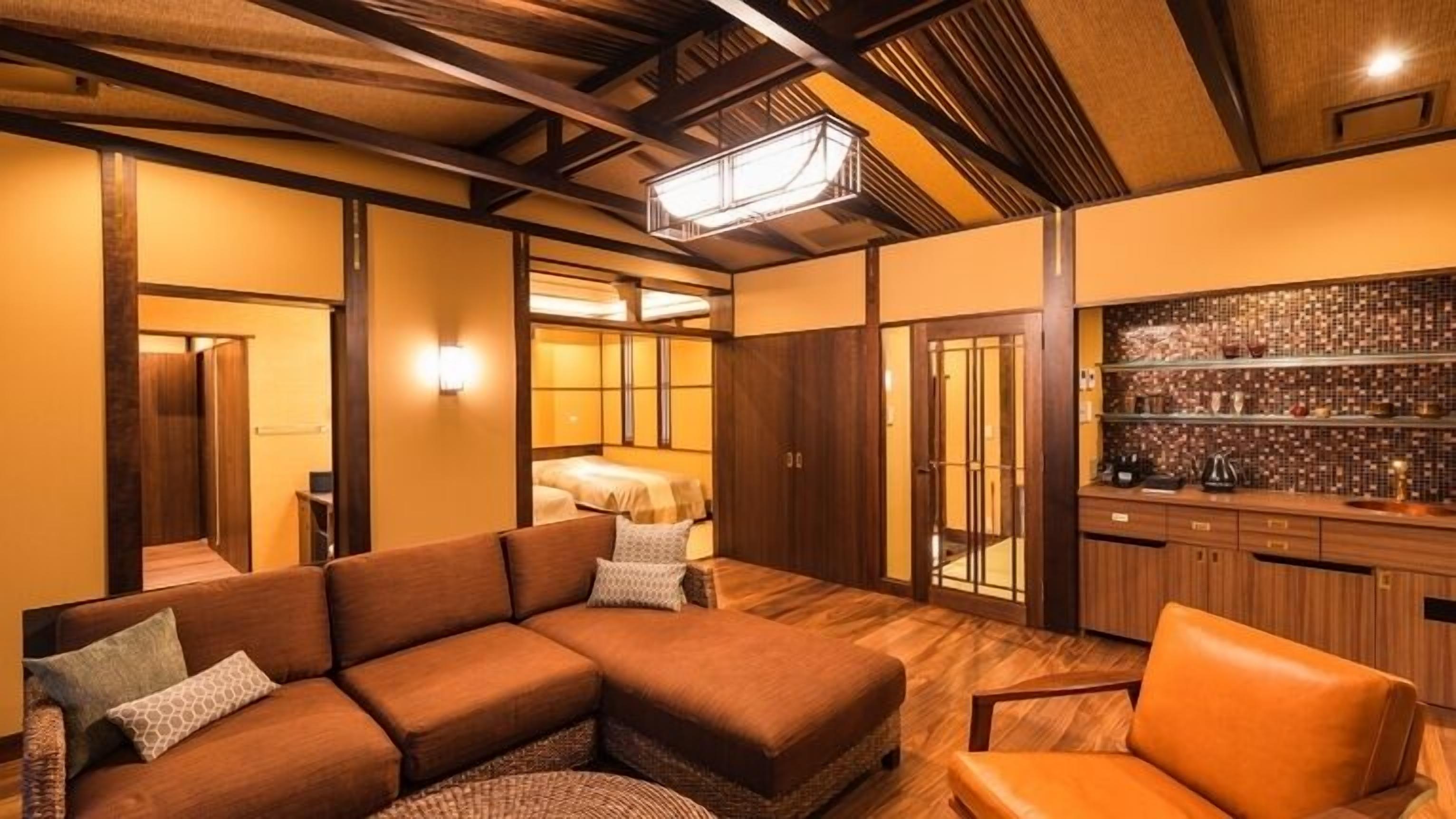 A hotel room with a sectional sofa, a single chair, a minibar, and a view of a bedroom.