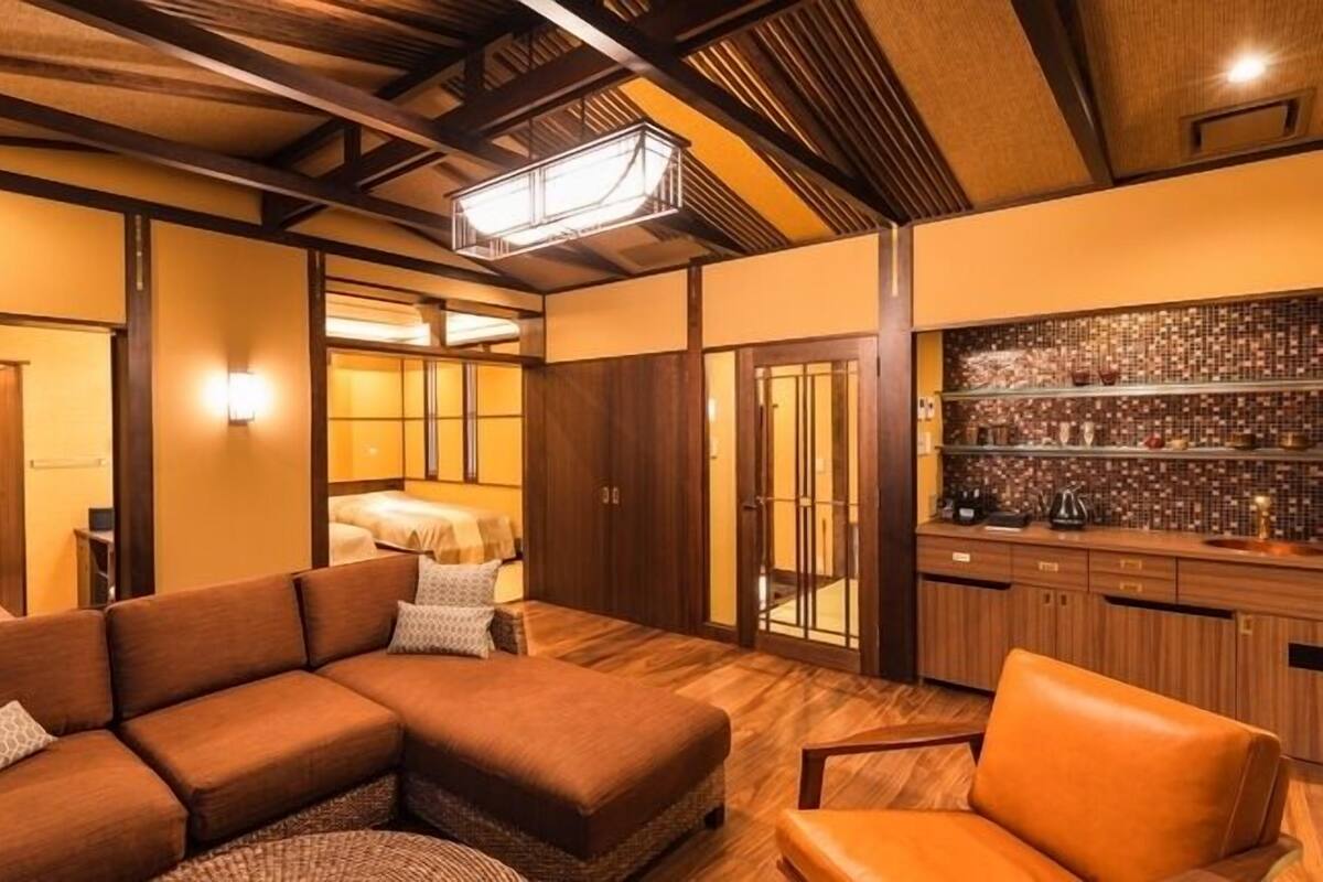 Habitación de hotel con sofá modular, una silla, minibar y vista a un dormitorio.