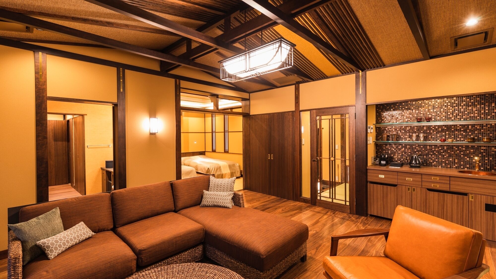 A modern hotel room with a sectional sofa, a small chair, a wooden floor, a kitchen area with a tiled backsplash, and a traditional Japanese-style sliding door.