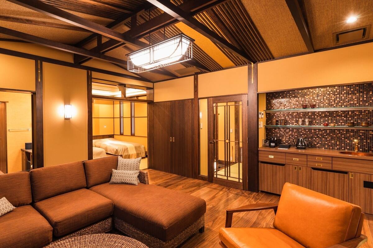 Una habitación de hotel moderna con sofá modular, una silla pequeña, piso de madera, área de cocina con salpicadero de azulejos y una puerta corredera de estilo japonés tradicional.