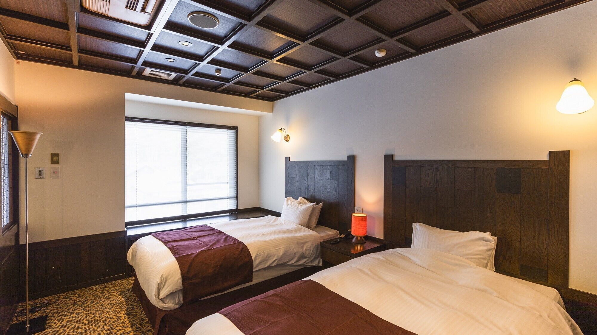 A hotel room with two beds, a window with blinds, a ceiling with recessed lighting, and a bedside table with a lamp.