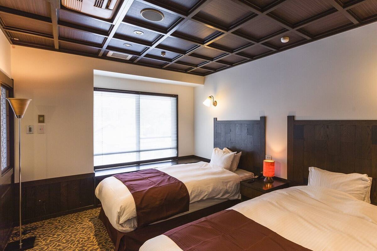 Habitación de hotel con dos camas, una ventana con persianas, techo con iluminación empotrada y una mesita de noche con lámpara.