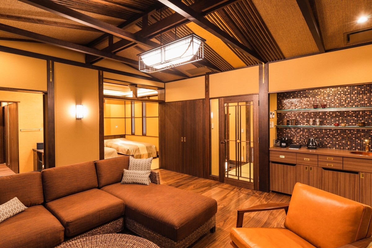 A modern hotel room with a sectional sofa, a small chair, a wooden floor, a kitchen area with a tiled backsplash, and a traditional Japanese-style sliding door.