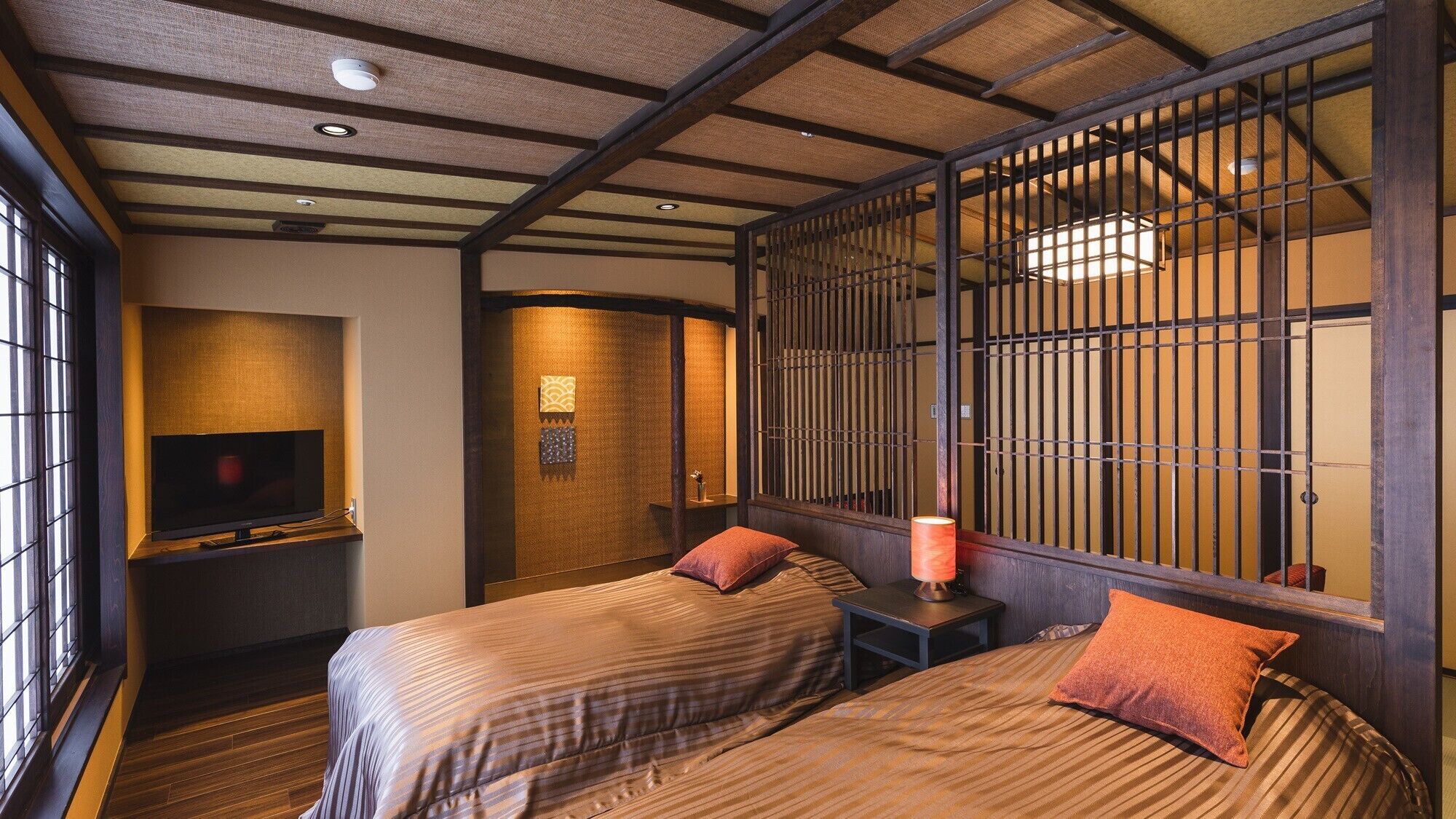 A traditional Japanese-style room with tatami flooring, a television, two beds with pillows, a sliding door with a grid pattern, and a small table with a lamp.