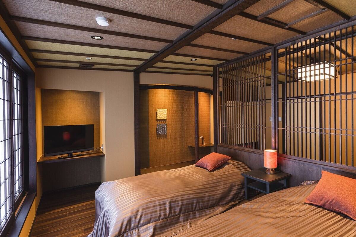 Una habitación de estilo japonés tradicional con suelo de tatami, un televisor, dos camas con almohadas, una puerta corredera con diseño de cuadrícula y una mesita con lámpara.