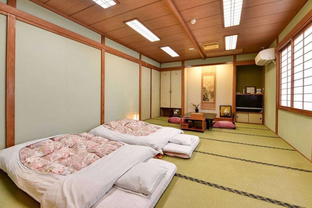 Une pièce de style japonais traditionnel, avec un sol recouvert de tatamis, deux lits avec une literie à motifs floraux, un futon, un bureau en bois et une porte coulissante.