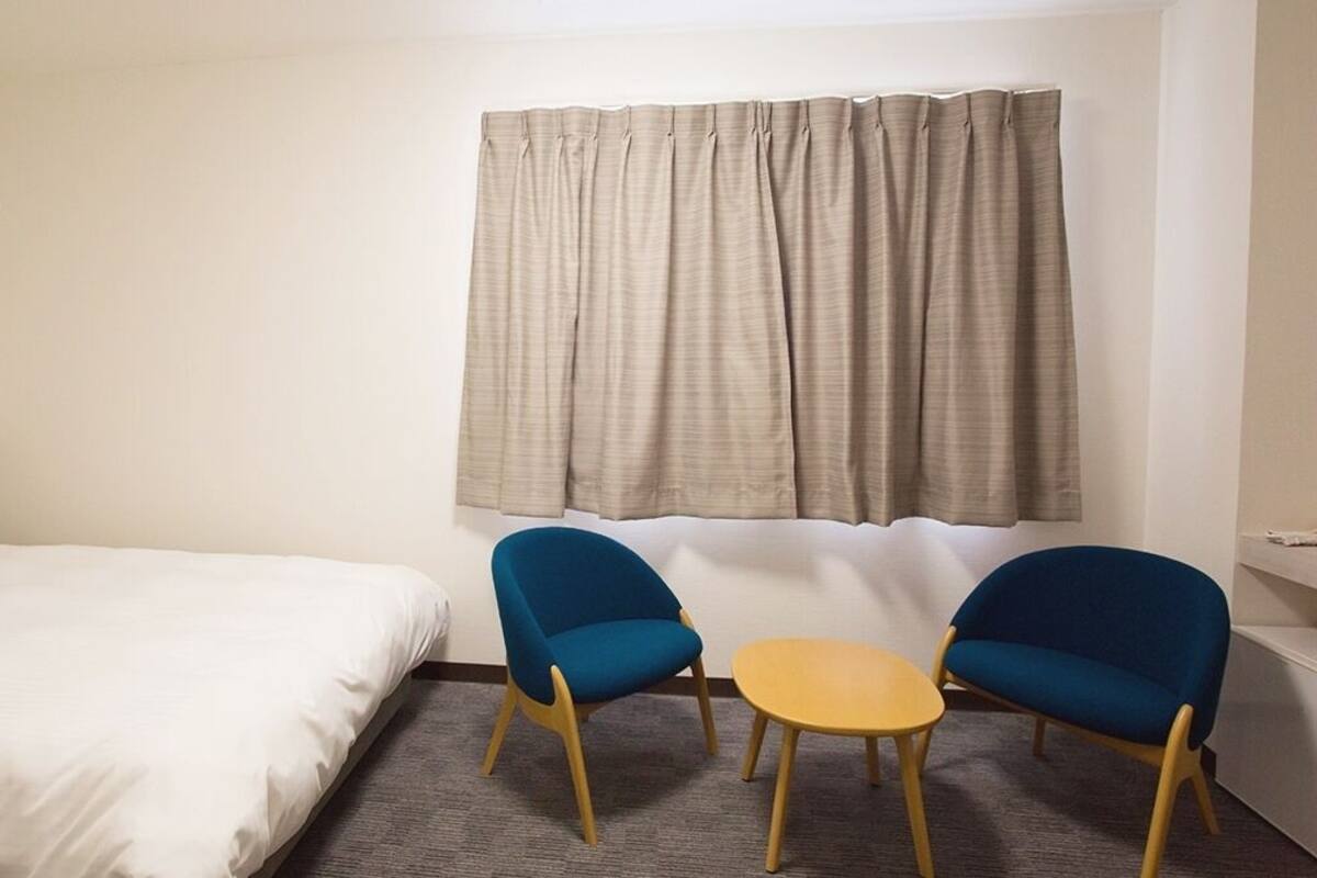 Une chambre d’hôtel avec un lit, deux chaises bleues, une petite table ronde et une fenêtre avec des rideaux.