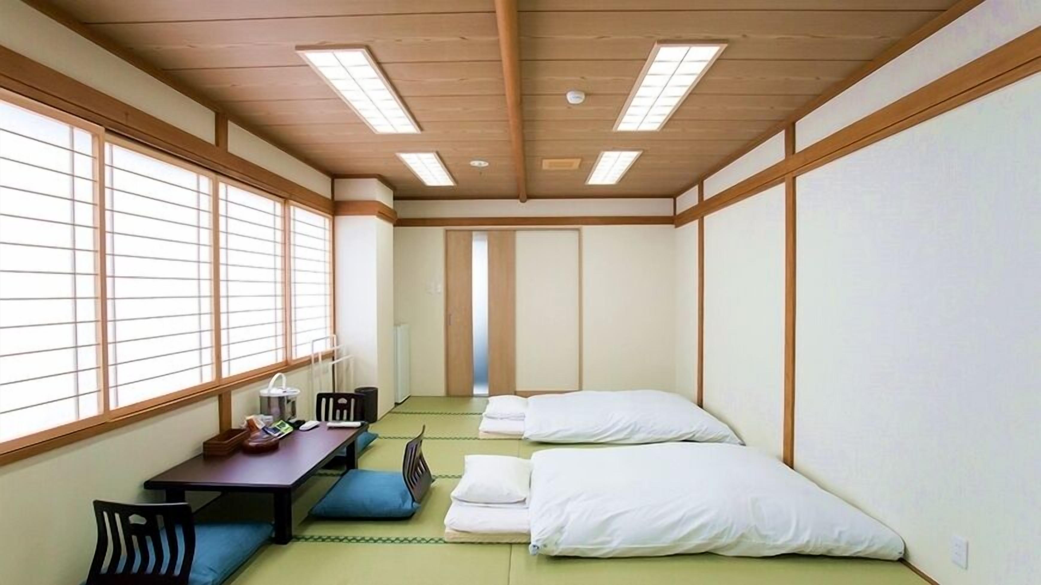 Een kamer met tatami vloerbedekking, een futonbed, een bureau met stoel en grote ramen met shoji schermen.