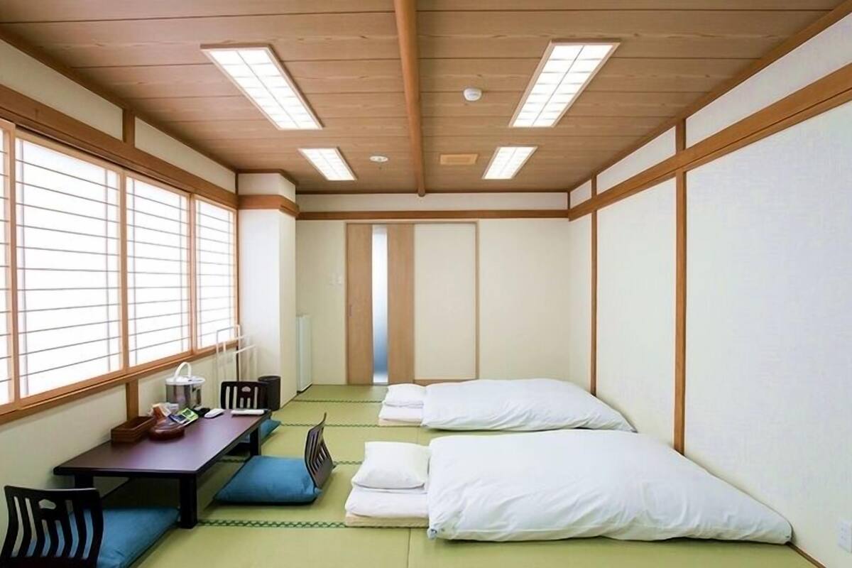 Une pièce avec un sol recouvert de tatamis, un futon servant de lit, un bureau avec une chaise et de grandes fenêtres équipées de paravents en papier de riz (shoji).